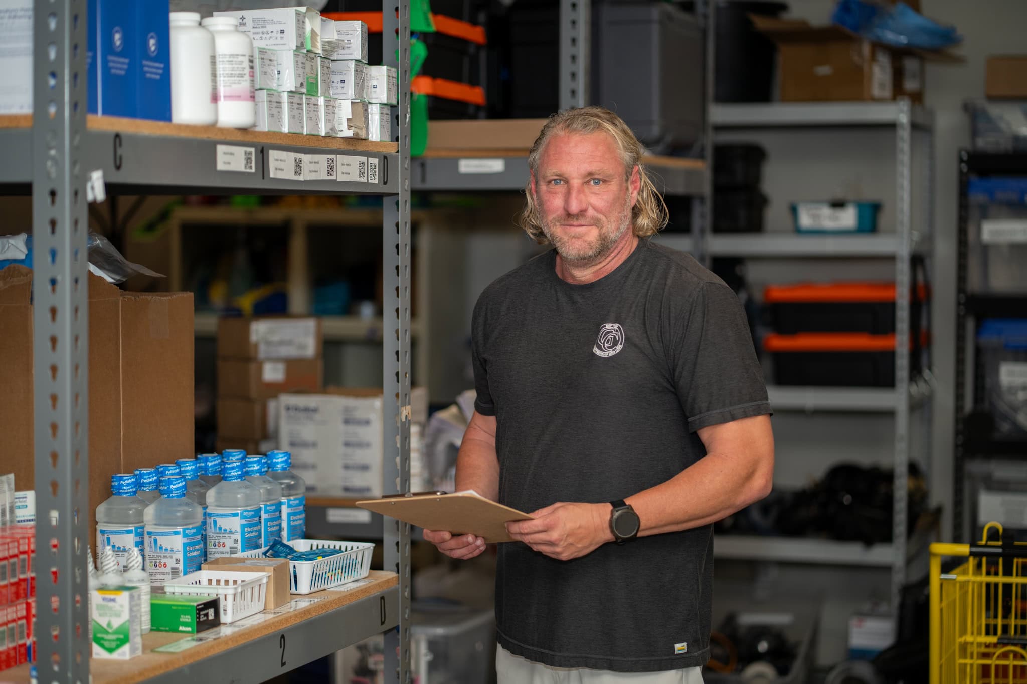 Handsome man with long blond hair holding a clipboard in a storage room filled with medical supplies, healthcare products, and equipment in the Gulf Coast region.