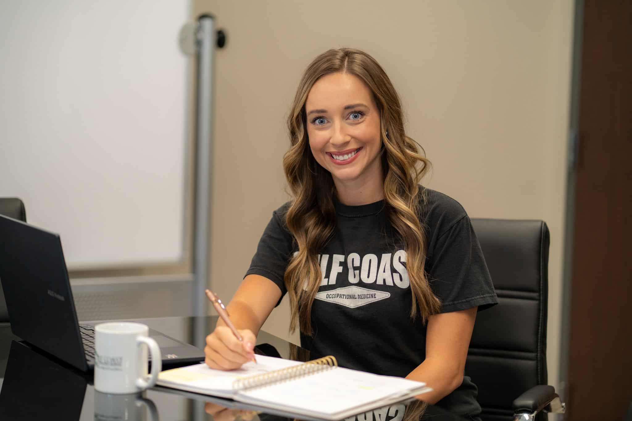 Professional woman in an office setting working at her desk, smiling, with a laptop and notepad, representing Gulf Coast occupational medicine and healthcare services.